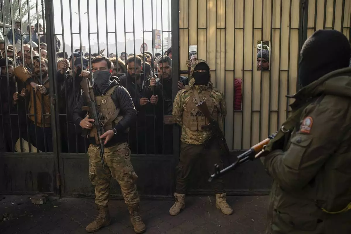 Syrian rebels stand in guard as members of Bashar Assad's army or a pro-government militia gather outside to be registered as part of an "identification and reconciliation process" in Damascus, Syria, Saturday, Dec. 21, 2024. (AP Photo/Leo Correa)