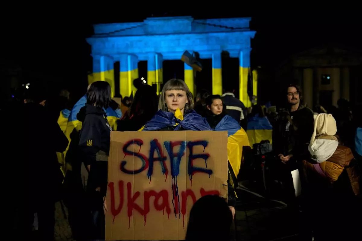 A protestor holds a placard at the Brandenburg Gate, illuminated in Ukrainian colors, during a demonstration against Russia's war on Ukraine as they mark the third anniversary of the full-scale invasion in Berlin, Monday, Feb. 24, 2025. (AP Photo/Ebrahim Noroozi)