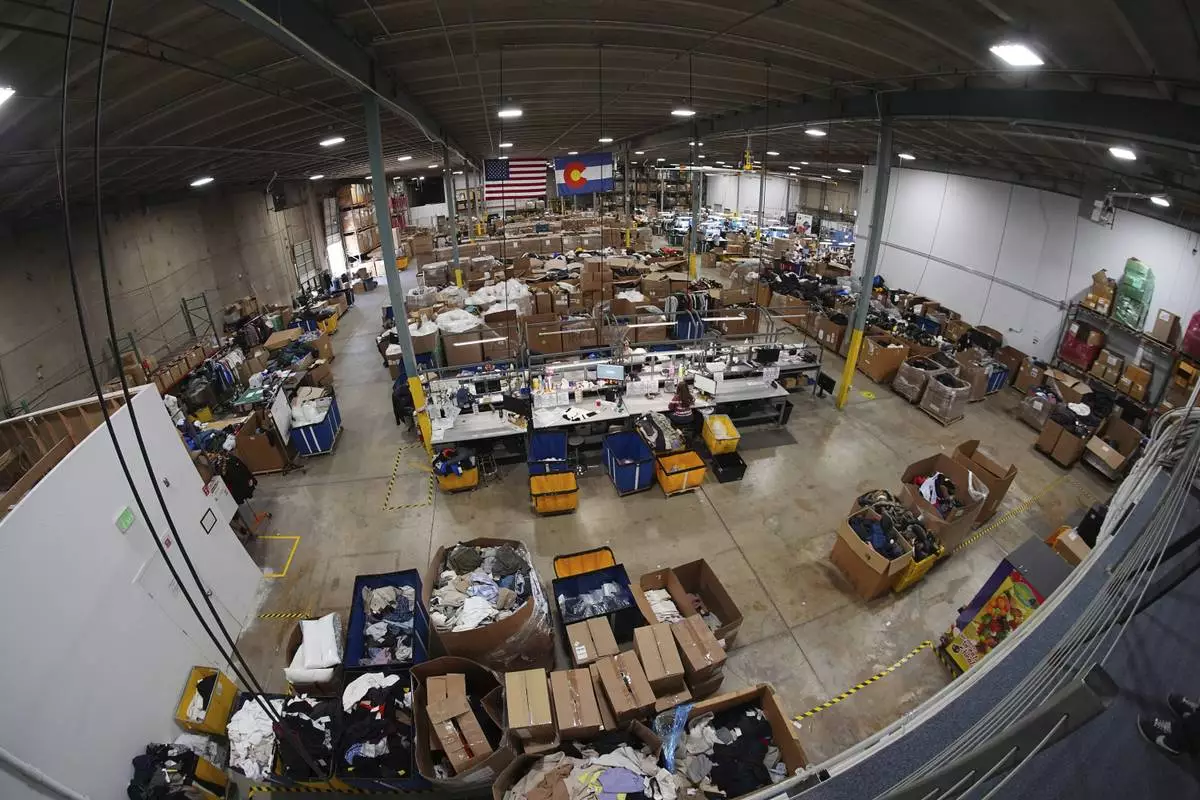 Seen through a fisheye lens, people work amid boxes of returned or overstocked clothing, shoes, boots, coats, packs and other items in a warehouse where the goods are cleaned or repaired before they are marketed on resale platforms in Englewood, Colo., on Thursday, April 17, 2025. (AP Photo/David Zalubowski)
