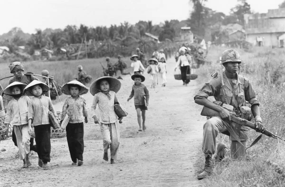 FILE- In an area heavily infiltrated by Viet Cong, a U.S. 1st Division soldier guards Route 7 as Vietnamese market women and schoolchildren return home to the village of Xuan Dien from Ben Cat, December 1965. (AP Photo/Horst Faas, File)