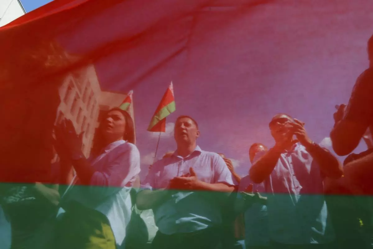 FILE – Belarusian government supporters, seen through a state flag, listen to a speaker at a rally at Independence Square in Minsk, Belarus, on Aug. 16, 2020. (AP Photo, File)