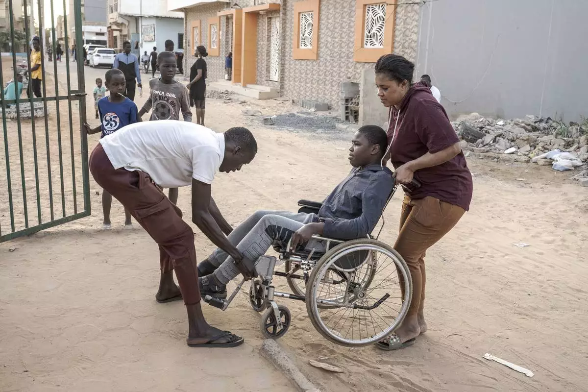 A man helps Adiaratou Ba, right, lift her son Mamadou, 13, suffering from a rare genetic disease, onto the curb in Dakar, Senegal, Saturday Jan. 11, 2025. (AP Photo/Annika Hammerschlag)
