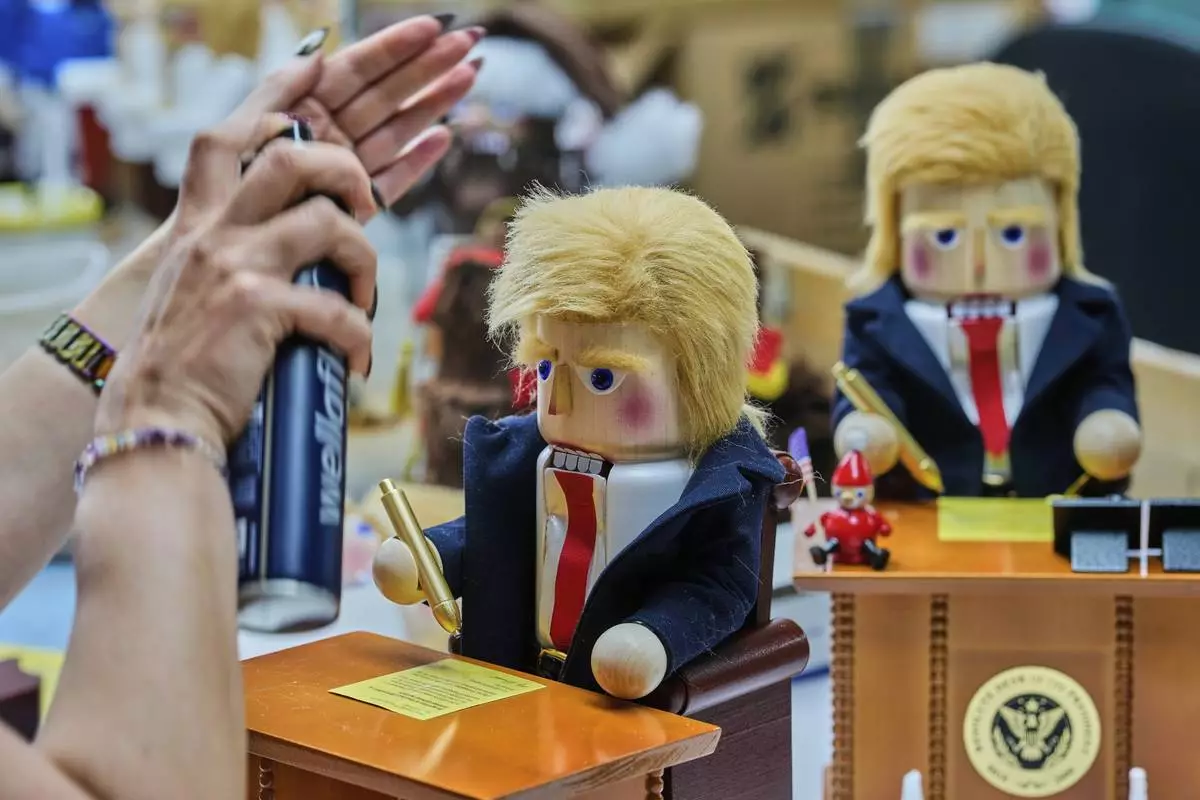 A woman uses hairspray to fixe the hair on a nutcracker titled "Resolute Desk of the President," featuring a seated figure signing a "presidential proclamation," at the Steinbach-Volkskunst nutcracker factory in Marienberg, Germany, Tuesday, April 15, 2025. (AP Photo/Markus Schreiber)