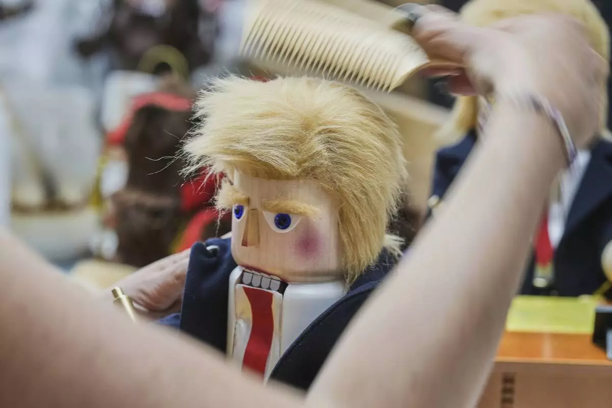 A woman combs the hair on a nutcracker titled "Resolute Desk of the President," featuring a seated figure signing a "presidential proclamation," at the Steinbach-Volkskunst nutcracker factory in Marienberg, Germany, Tuesday, April 15, 2025. (AP Photo/Markus Schreiber)