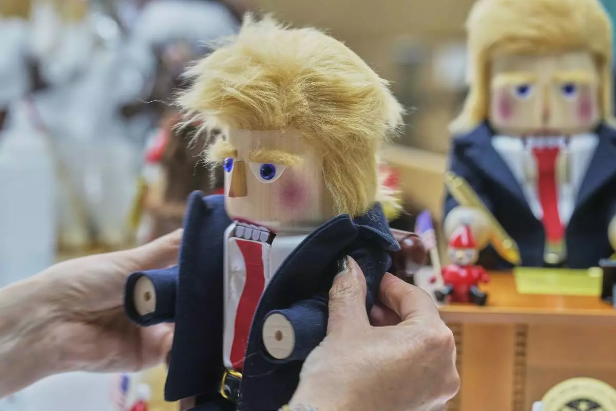 A woman fixes a coat on a nutcracker titled "Resolute Desk of the President," featuring a seated figure signing a "presidential proclamation," at the Steinbach-Volkskunst nutcracker factory in Marienberg, Germany, Tuesday, April 15, 2025. (AP Photo/Markus Schreiber)