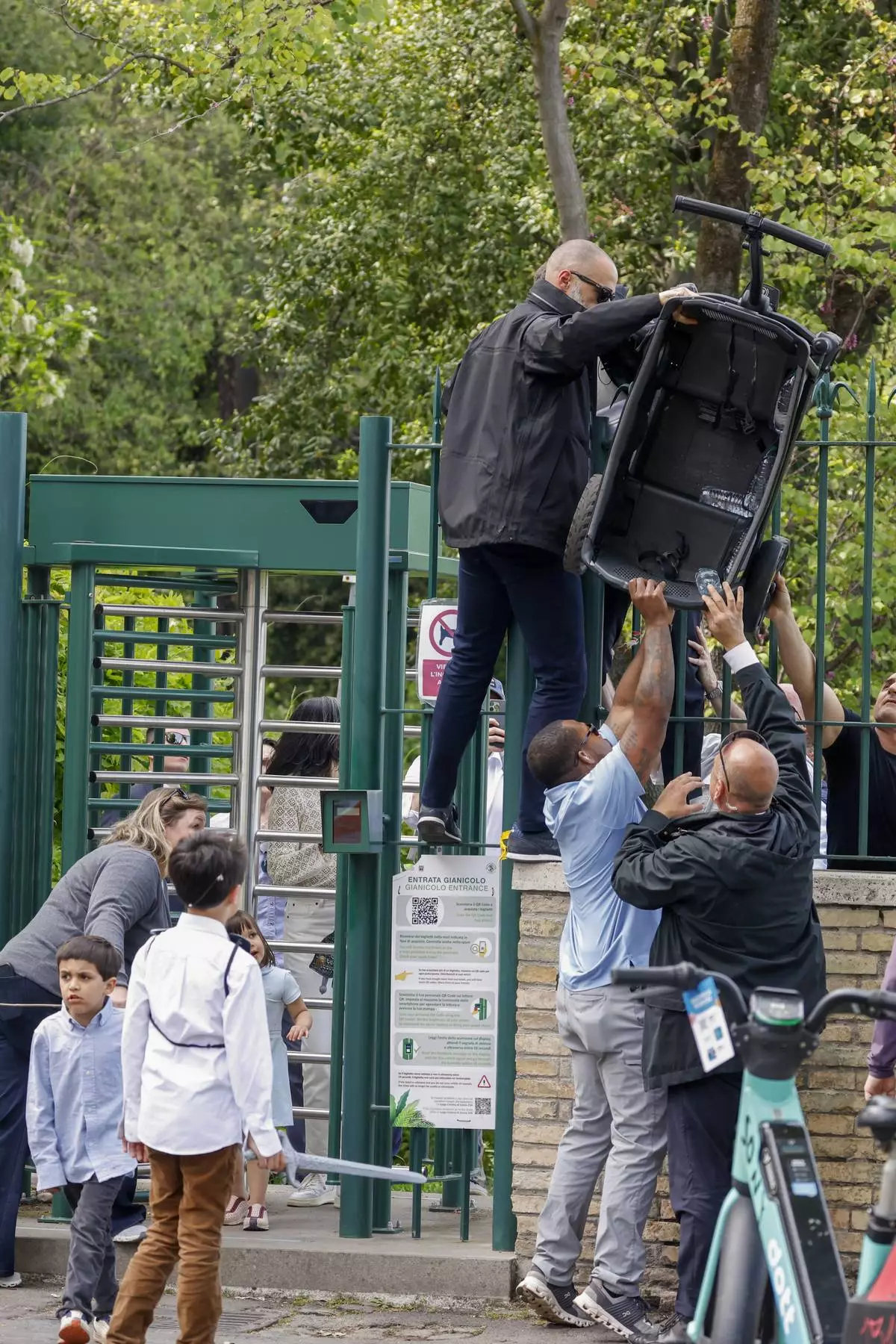 Security personnel accompanying U.S. Vice President JD Vance lift a trolley over the turnstiles at the exit of Rome's Botanic Gardens after a tour inside the gardens Vance did with his wife Usha, partially hidden behind the gate, their daughter Mirabel, sons Vivek, left, and Ewan, back to camera, Saturday, April 19, 2025. (AP Photo/Riccardo De Luca)