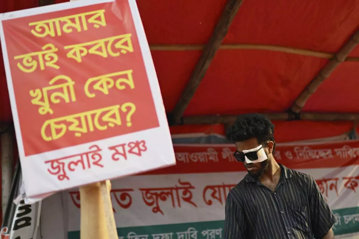 A man who was injured in protests against former Prime Minister Sheikh Hasina last year, stands next to a banner that reads 'My brother is in the grave, why is the murderer in the chair?' during a protest to demand a ban on her Awami League party, in Dhaka, Bangladesh, Sunday, May 11, 2025. (AP Photo/Mahmud Hossain Opu)