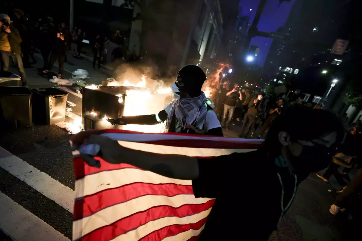 A protester holds up an American flag in front of a dumpster that was set on fire during a protest against federal immigration arrests, Wednesday, June 11, 2025, in Seattle. (AP Photo/Ryan Sun)