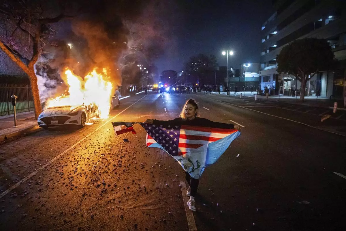 A person carrying multiple flags walks past a burning car during protests over the Trump administration's immigration raids in Los Angeles, Monday, June 9, 2025. (AP Photo/Ethan Swope)