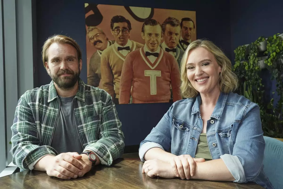 Chris Candy, left, and Jennifer Candy-Sullivan, the children of the late John Candy, pose in front of a photograph of The Second City comedy performers including their father, far left, to promote the documentary film "John Candy: I Like Me" during the Toronto International Film Festival on Thursday, Sept. 4, 2025, in Toronto. (AP Photo/Chris Pizzello)