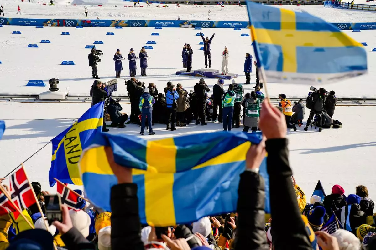Sweden fans wave flags as Frida Karlsson, of Sweden, jumps on the podium celebrating after winning the gold medal in the cross country skiing women's 10km interval start free at the 2026 Winter Olympics, in Tesero, Italy, Thursday, Feb. 12, 2026. (AP Photo/Evgeniy Maloletka)
