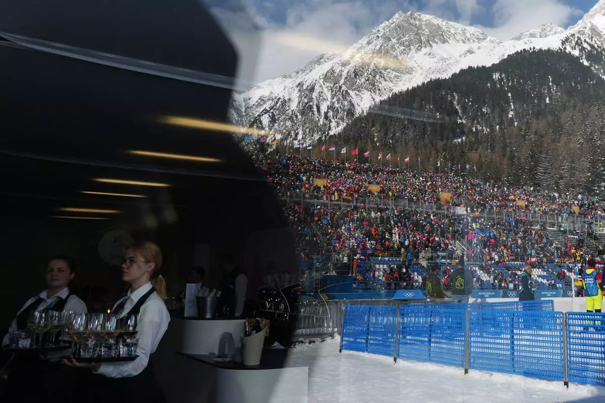 Waiters serve drinks as fans are reflected on a building ahead of the men's 15-kilometer mass start biathlon race at the 2026 Winter Olympics in Anterselva, Italy, Friday, Feb. 20, 2026. (AP Photo/Andrew Medichini)