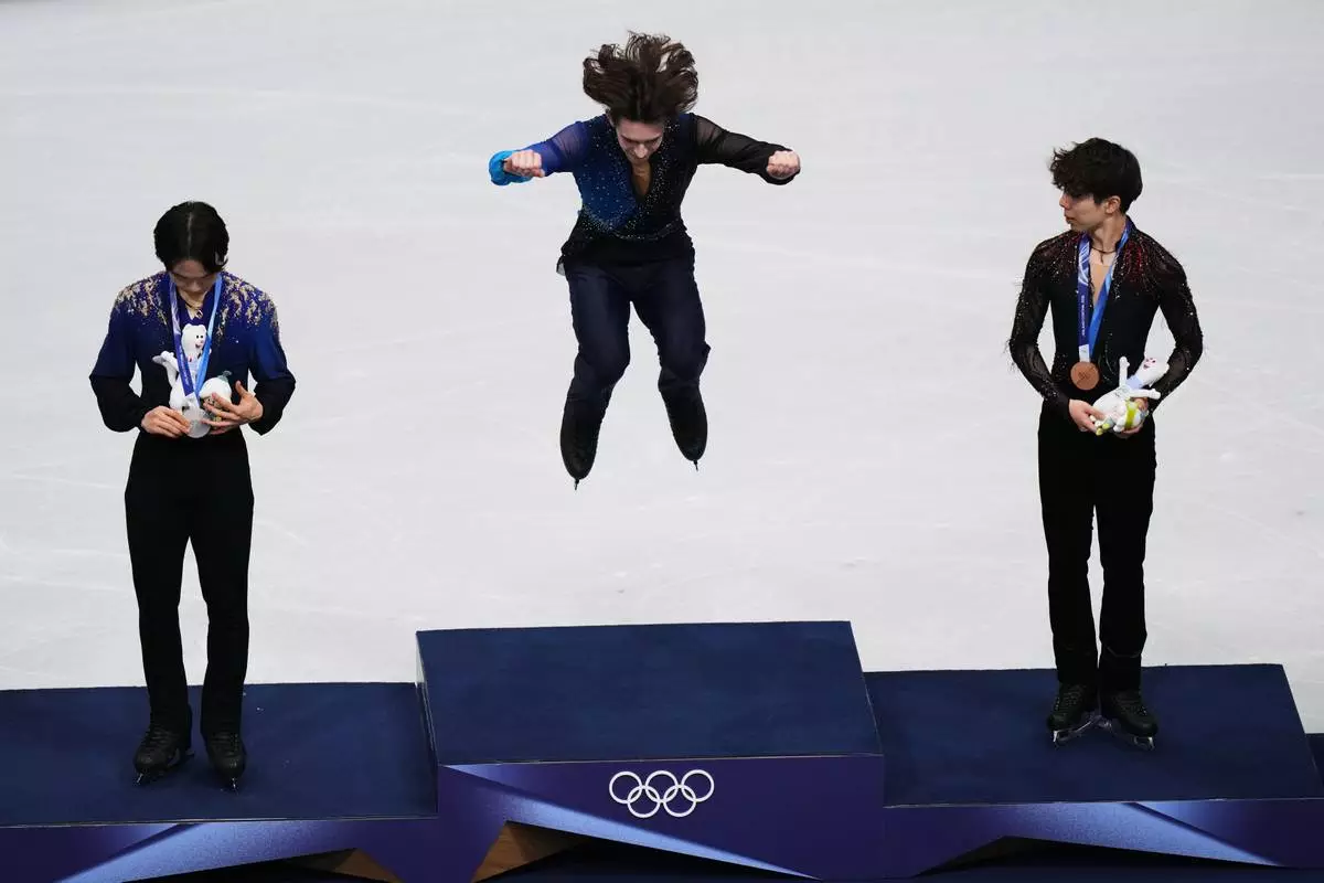 Gold medalist Mikhail Shaidorov of Kazakhstan jumps up to the podium to receive his medal with silver medalist Yuma Kagiyama of Japan, left, and bronze medalist Shun Sato of Japan, right, after competing in the men's free skate program in figure skating at the 2026 Winter Olympics, in Milan, Italy, Friday, Feb. 13, 2026. (AP Photo/Francisco Seco)