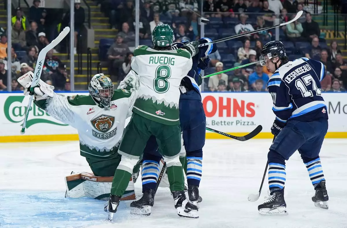 The puck deflects off of Chicoutimi Sagueneens' Christophe Berthelot, back centre, as he's checked by Everett Silvertips' Kayd Ruedig (8) in front of goalie Anders Miller (31) as Chicoutimi's Thomas Desruisseaux (17) watches during the second period of a Memorial Cup hockey game in Kelowna, British Columbia, Saturday, May 23, 2026. (Darryl Dyck/The Canadian Press via AP)