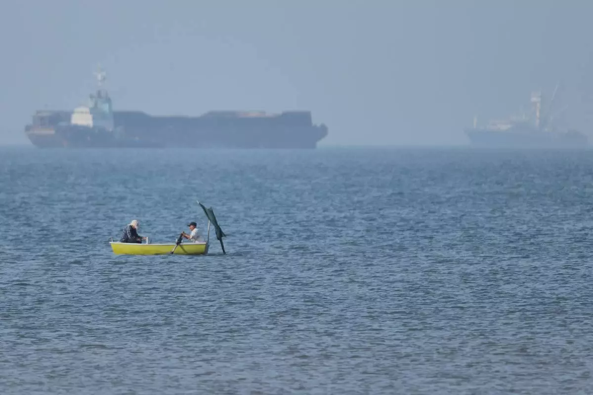 Two men sit in a small boat on the water as cargo ships are anchored in the background in the Strait of Hormuz off Bandar Abbas, Iran, Monday, May 4, 2026. (Amirhosein Khorgooi/ISNA via AP)