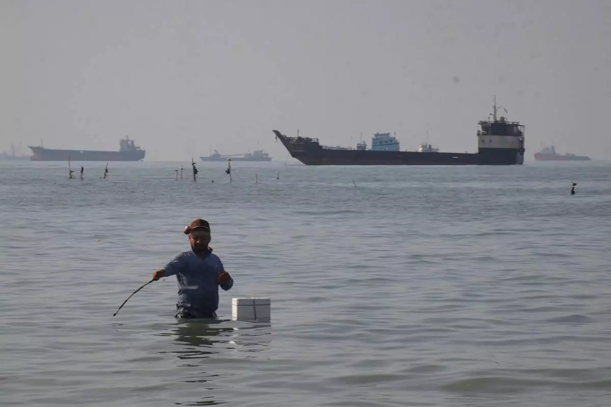 A man stands in the water, appearing to fish, as bulk carriers, cargo ships, and service vessels line the horizon in the Strait of Hormuz off Bandar Abbas, Iran, Monday, April 27, 2026.(Razieh Poudat/ISNA via AP)