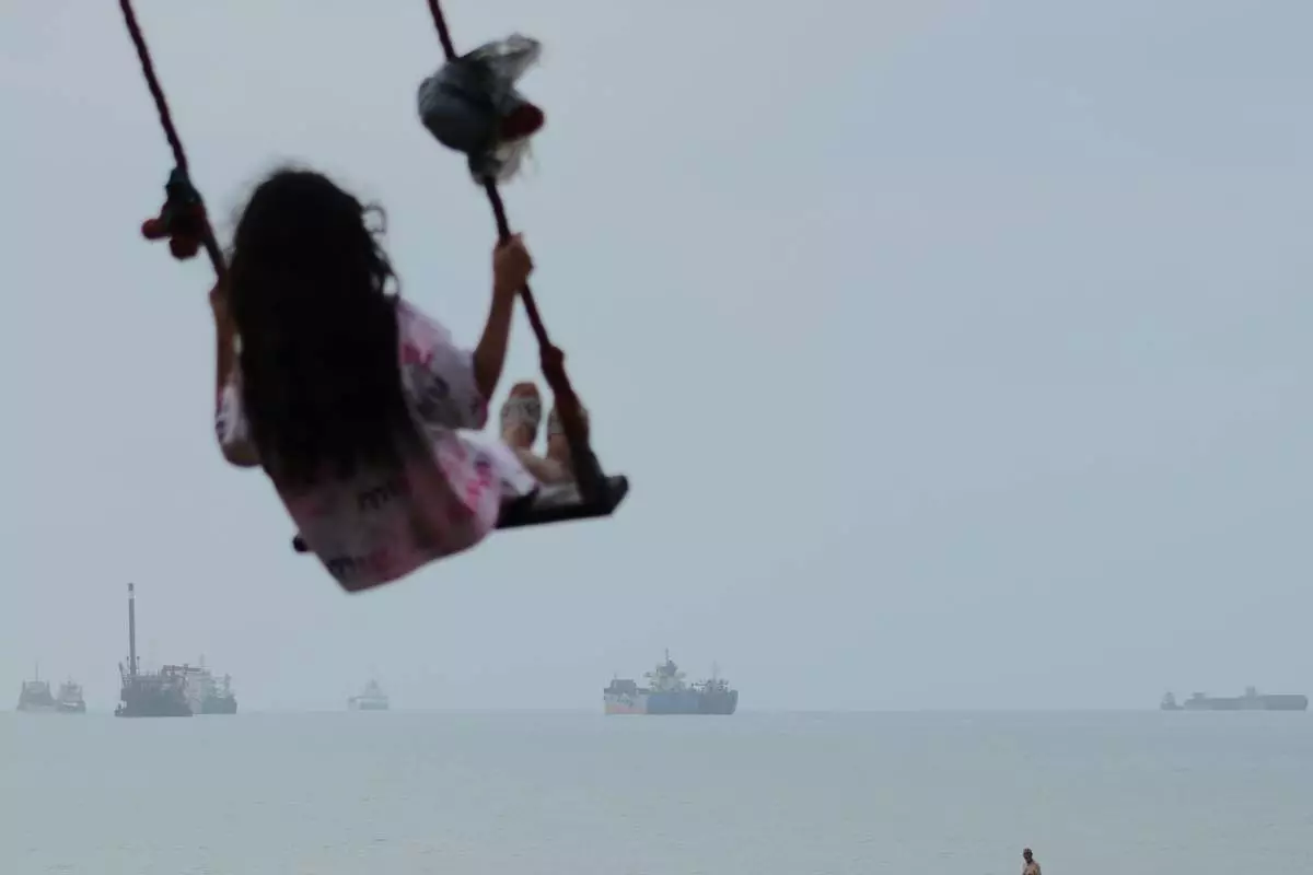 A girl swings along the shore as cargo ships and other commercial vessels appear anchored in the Strait of Hormuz off Bandar Abbas, Iran, Monday, July 27, 2026. (Razieh Poudat/ISNA via AP)