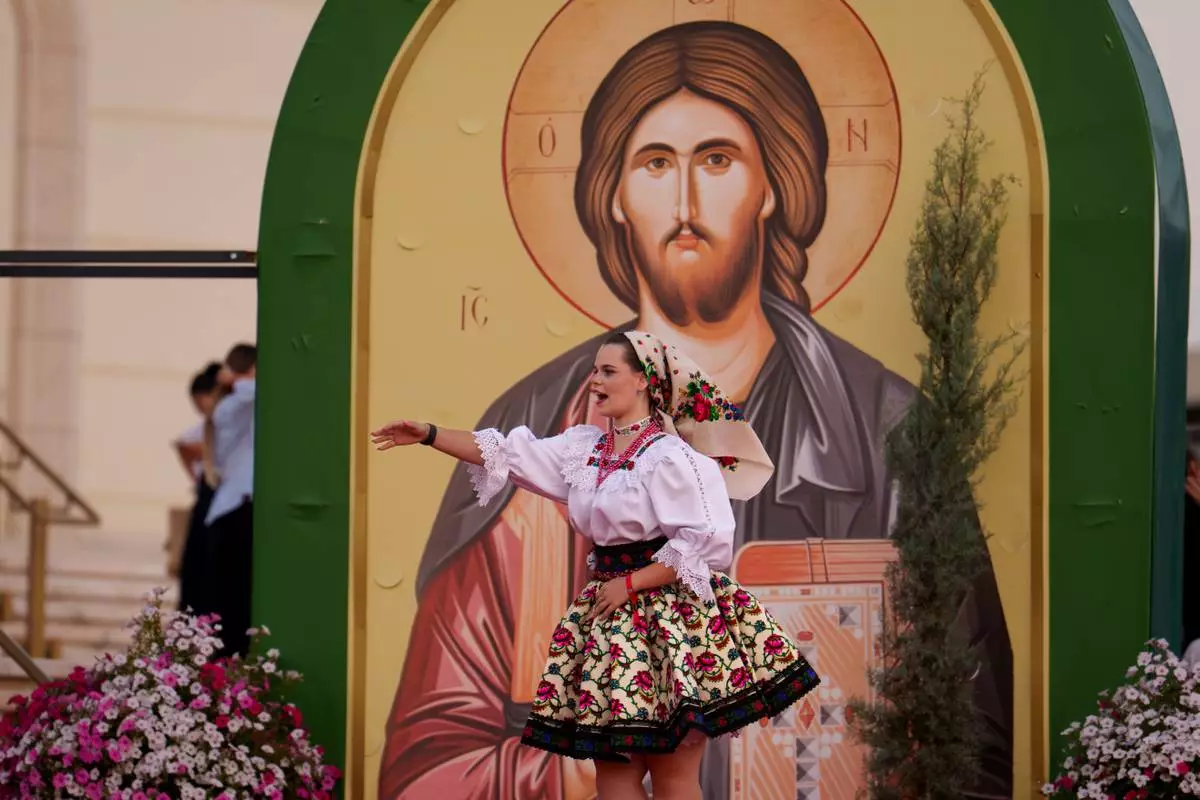 A young woman wearing a traditional outfit gestures backdropped by a religious icon before the Procession of Lights, part of a four-day program of the International Meeting of Orthodox Youth (ITO), in Bucharest, Romania, Wednesday, Sept. 2, 2026. (AP Photo/Andreea Alexandru)