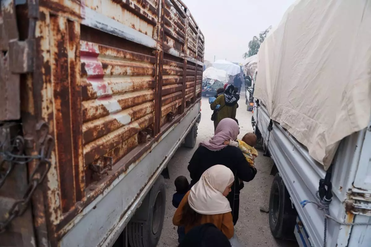 Women carry young children between trucks loaded with belongings as displaced families prepare to leave al-Talayi camp in Hassakeh, Syria, Tuesday, Oct. 13, 2026. Around 450 families are returning to the Ras al-Ayn countryside, seven years after a Turkish military offensive forced them to flee. (AP Photo/Baderkhan Ahmad)