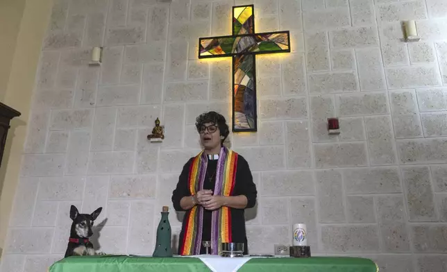 Rev. Elaine Saralegui, wearing a rainbow-colored clergy stole and her clerical collar, welcomes congregants to a service at the Metropolitan Community Church, an LGBTQ+ inclusive house of worship, as Ruth the dog stands with her front paws on the altar table, in Matanzas, Cuba, Friday, Feb. 2, 2024. In 2015, with support from the U.S.-based LGBTQ+ affirming Metropolitan Community Churches, they converted a house into their church, decked with wooden pews and a stained-glass cross that hangs above the altar. (AP Photo/Ramon Espinosa)
