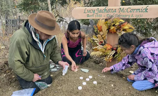 In this Sept. 29, 2023, photo at the grave of Lucky Pitka McCormick, her granddaughter Kathleen Carlo, left, and McCormick's great-great-grandchildren Lucia, center, and Addison Carlo place candles and stones on the grave during a reburial ceremony in Rampart, Alaska. Pitka was one of the Lost Alaskans sent to a mental hospital in the 1930s. Her grave was recently discovered, and family members brought her back to Alaska for a proper burial. (Wally Carlo via AP).