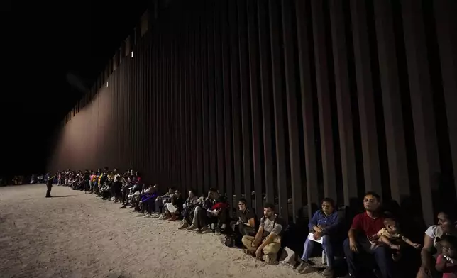 FILE - Migrants wait along a border wall Tuesday, Aug. 23, 2022, after crossing from Mexico near Yuma, Ariz. The UN migration agency marks a decade since the launch of the Missing Migrants Project, documenting more than 63,000 deaths around the world. More than two-thirds of victims remain unidentified highlighting the size of the crisis and the suffering of families who rarely receive definitive answers. (AP Photo/Gregory Bull, File)