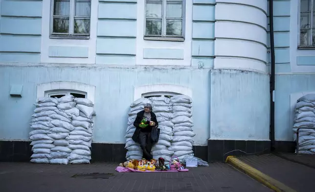 A woman sells toys in front of a building with windows protected by sandbags in Kyiv, Ukraine, Sunday, April 28, 2024. (AP Photo/Francisco Seco)