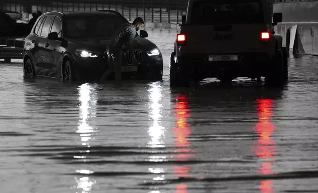 A man tries to work on his stalled SUV in standing water in Dubai, United Arab Emirates, Tuesday, April 16, 2024. Heavy rains lashed the United Arab Emirates on Tuesday, flooding out portions of major highways and leaving vehicles abandoned on roadways across Dubai. Meanwhile, the death toll in separate heavy flooding in neighboring Oman rose to 18 with others still missing as the sultanate prepared for the storm. (AP Photo/Jon Gambrell)
