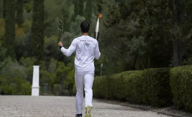 The first torch bearer, Greek olympic gold medalist Stefanos Douskos, runs towards the monument to Pierre de Coubertin, in the background, after the official ceremony of the flame lighting for the Paris Olympics, at the Ancient Olympia site, Greece, Tuesday, April 16, 2024. The flame will be carried through Greece for 11 days before being handed over to Paris organizers on April 26. (AP Photo/Thanassis Stavrakis)