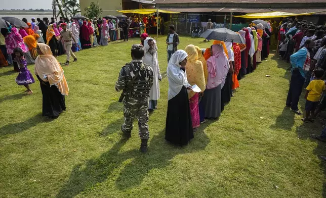 A security person walks past queues of voters in a polling station on the bank of the Brahmaputra river during the second round of voting in the six-week-long national election in Morigaon district, Assam, India, Friday, April 26, 2024. (AP Photo/Anupam Nath)