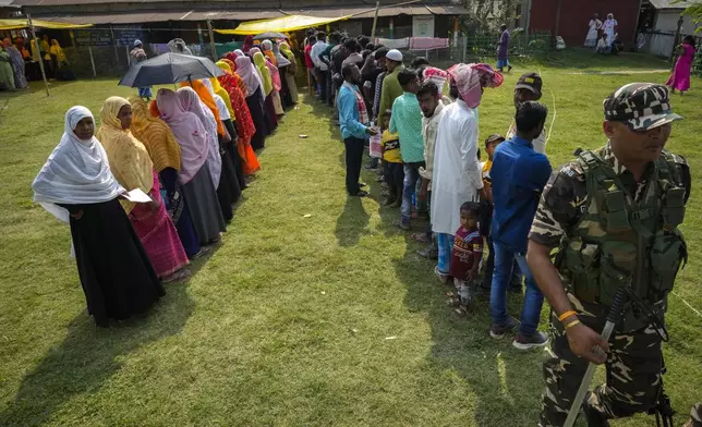 A security person walks past queues of voters in a polling station on the bank of the Brahmaputra river during the second round of voting in the six-week-long national election in Morigaon district, Assam, India, Friday, April 26, 2024. (AP Photo/Anupam Nath)