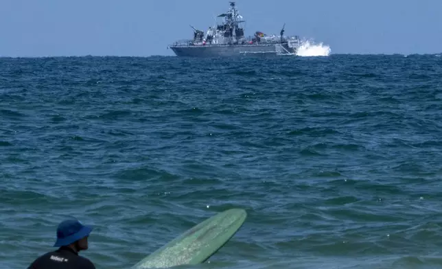 A surfer waits for wave while an Israeli military naval ship patrols the Mediterranean sea off the coast of Hadera, Israel, Sunday, April 14, 2024. Israel on Sunday hailed its successful air defenses in the face of an unprecedented attack by Iran, saying it and its allies thwarted 99% of the more than 300 drones and missiles launched toward its territory. But regional tensions remain high, amid fears of further escalation in the event of a possible Israeli counter-strike. (AP Photo/Ariel Schalit)