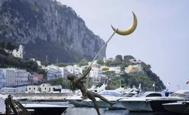 The bronze sculpture by Giacinto Bosco titled "swing on the moon" is backdropped by Capri's harbour, Wednesday, April 17, 2024. Group of Seven foreign ministers are meeting on the Italian resort island of Capri, with soaring tensions in the Mideast and Russia's continuing war in Ukraine topping the agenda. The meeting runs April 17-19. (AP Photo/Gregorio Borgia)