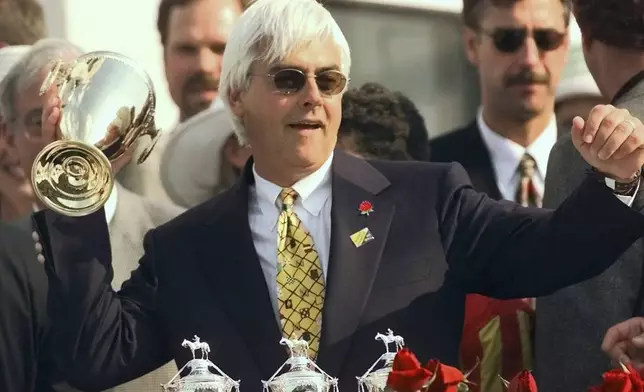 FILE - Real Quiet trainer Bob Baffert strikes a quarterback pose with the Kentucky Derby trophy after Real Quiet with jockey Kent Desormeaux up won the 124th running of the Kentucky Derby at Churchill Downs, in Louisville, Ky., May 2, 1998. Baffert will miss the race for the third consecutive year. He served a two-year suspension by Churchill Downs Inc. after his 2021 winner Medina Spirit was disqualified for a failed drug test. But the track’s corporate ownership meted out an additional year of punishment. (AP Photo/Ed Reinke, File)