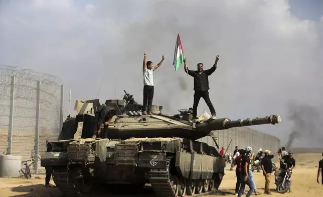 FILE - Palestinians wave their national flag and celebrate by a destroyed Israeli tank at the southern Gaza Strip fence east of Khan Younis on Saturday, Oct. 7, 2023. Thousands of Hamas-led militants stormed across the border into Israel, killing 1,200 people, mostly civilians, and taking roughly 250 captive, according to Israeli authorities. (AP Photo/Yousef Masoud, File)