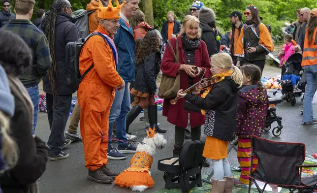 An orange-clad man and his orange-clad dog listen to a girl playing violin during King's Day celebrations in Amsterdam, Netherlands, Saturday, April 27, 2024. (AP Photo/Peter Dejong)