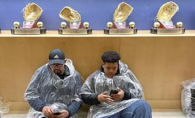 Fans wait in a lobby under Gold Gloves during a rain delay before a baseball game between the Los Angeles Dodgers and the San Diego Padres, Saturday, April 13, 2024, in Los Angeles. (AP Photo/Marcio Jose Sanchez)