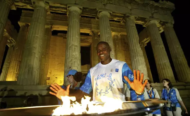 FILE - Nine times Olympic gold medal winner Carl Lewis of the United States poses for the photographers in front of the Olympic flame in front of the columns of the ancient Parthenon temple at the Acropolis hill, Aug. 12, 2004. On Tuesday, April 16, 2024 the flame for this summer's Paris Olympics will be lit and be carried through Greece for more than 5,000 kilometers (3,100 miles) before being handed over to French organizers at the Athens site of the first modern Olympics. (AP Photo/Lefteris Pitarakis, File)