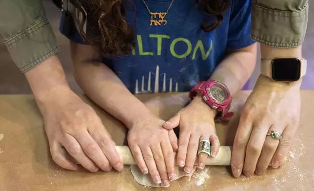 Wearing a necklace with her name in Hebrew, Charlotte Gleicher, 7, a first grader at Milton Gottesman Jewish Day School of the Nation's Capital, is helped by her teacher, Dafna Kiverstein, to roll out dough for matzah during a "Matzah Factory" field trip at the JCrafts Center for Jewish Life and Tradition in Rockville, Md., Thursday, April 18, 2024, ahead of the Passover holiday. To be kosher for Passover, which begins next Monday evening, the dough has to be prepared and cooked all within 18 minutes and not allowed to rise. (AP Photo/Jacquelyn Martin)