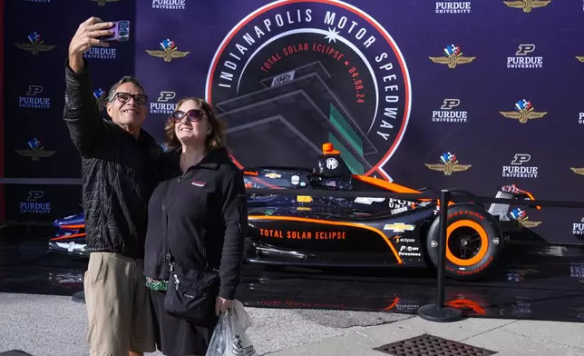 Vicki and Craig Musser, of Vernon Hills, Ill. take a selfie with a total eclipse themed race car at the Indianapolis Motor Speedway in Indianapolis, Monday, April 8, 2024. (AP Photo/Michael Conroy)