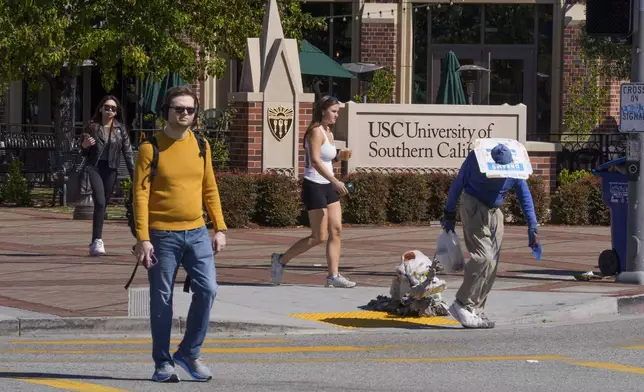 A person suffering homelessness walks past students outside the University of Southern California campus entrance in Los Angeles on Tuesday, April 16, 2024. University of Southern California officials have canceled a commencement speech by its 2024 valedictorian, a pro-Palestinian Muslim, citing "substantial risks relating to security and disruption" of the event that draws 65,000 people to campus. (AP Photo/Damian Dovarganes)