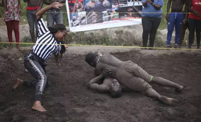 Ugandan youths perform an amateur wrestling tangle in the soft mud in Kampala, Uganda Wednesday, March. 20, 2023. The open-air training sessions, complete with an announcer and a referee, imitate the pro wrestling contests the youth regularly see on television. While a pair tangles inside the ring, made with bamboo poles strung with sisal rope, others standing ringside cheer feints and muscular shows of strength. (AP Photo/Patrick Onen)