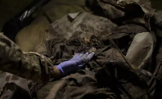 Oleksii Yukov inspects the necklace of a deceased Russian soldier in Sloviansk region, Ukraine, Tuesday, Oct. 24, 2023. “We are not fighting the dead,” he said. “I don’t separate the bodies of Russian soldiers and Ukrainian soldiers. They are all souls for me.” (AP Photo/Bram Janssen)