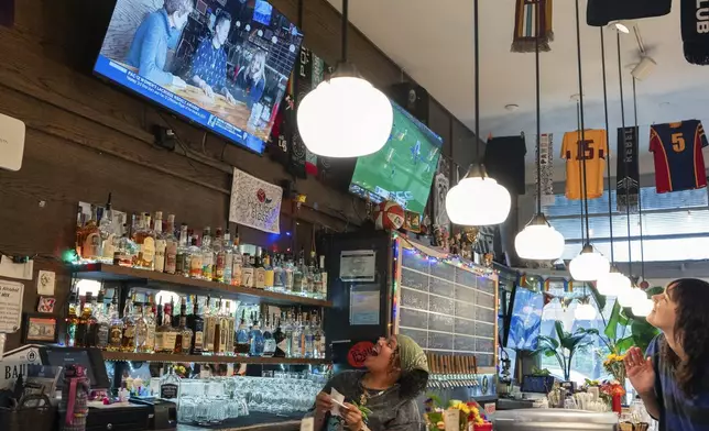 Bartender M.J. Jones, left, and fellow employee Allison Clarke, right, react as an interview with The Sports Bra founder and CEO Jenny Nguyen, center, airs on a screen during their shift at the sports bar on Wednesday, April 24, 2024, in Portland, Ore. A painting of U.S. soccer legend Abby Wambach is mounted above the chalkboard beer menu. (AP Photo/Jenny Kane)