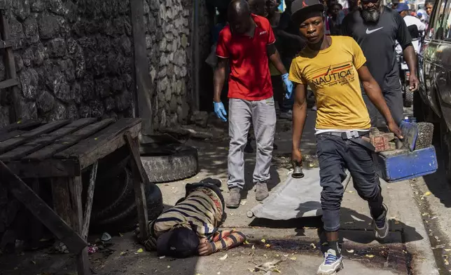 A shoeshiner walks past a bloodied body being removed by forensic workers in Port-au-Prince, Haiti, Wednesday, May 1, 2024. Forensics said the body had gunshot wounds. (AP Photo/Ramon Espinosa)