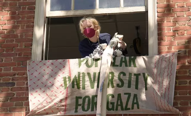 An activist uses keffiyeh scarves tied together to hoist a banner through an upper floor window of a Rhode Island School of Design building they have partially taken over Tuesday, May 7, 2024, at RISD, in Providence, R.I. The students and supporters are demanding that RISD condemn Israel's war effort in Gaza, and that the school divest from investments that benefit Israel. (AP Photo/Steven Senne)