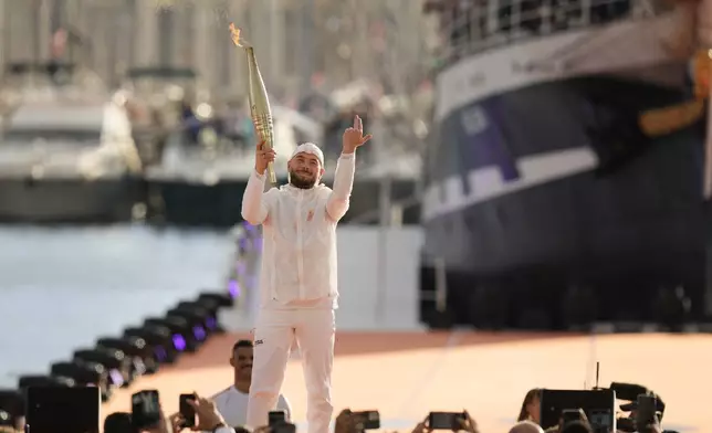 French rap artist Jul holds the Olympic torch during the torch arrival ceremony in Marseille, southern France, Wednesday May 8, 2024. After leaving Marseille, a vast relay route is undertaken before the torch odyssey ends on July 27 in Paris. The Paris 2024 Olympic Games will run from July 26 to Aug.11, 2024. (AP Photo/Thibault Camus)