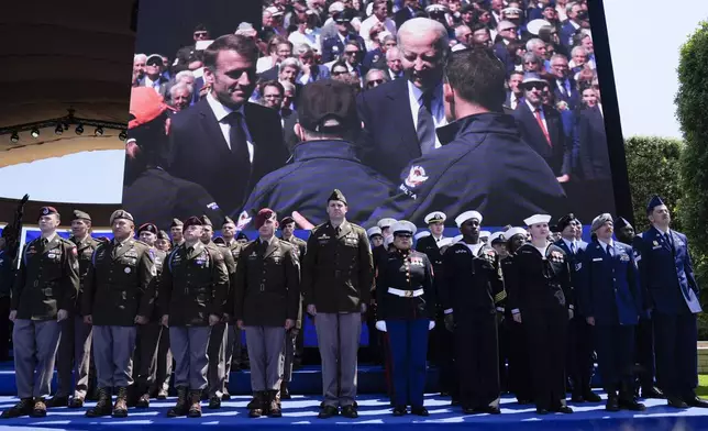 President Joe Biden and French President Emmanuel Macron are shown on a large screen greeting a World War II veteran during ceremonies to mark the 80th anniversary of D-Day, Thursday, June 6, 2024, in Normandy. (AP Photo/Evan Vucci)