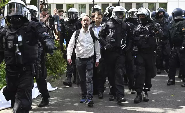 Participants of the AfD federal party conference are escorted by the police past demonstrators to the Grugahalle, in Essen, Germany, Saturday, June 29, 2024. The two-day national party conference of the AfD is taking place in the Grugahalle, including the election of the federal executive committee. Numerous organizations have announced opposition to the meeting and more than a dozen counter-demonstrations. (Henning Kaiser/dpa via AP)