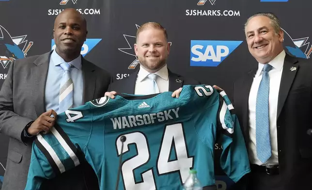 Ryan Warsofsky, middle, holds up a jersey between San Jose Sharks general manager Mike Grier, left, and president Jonathan Becher at a news conference introducing Warsofsky as the NHL hockey head coach of the Sharks in San Jose, Calif., Monday, June 17, 2024. (AP Photo/Jeff Chiu)